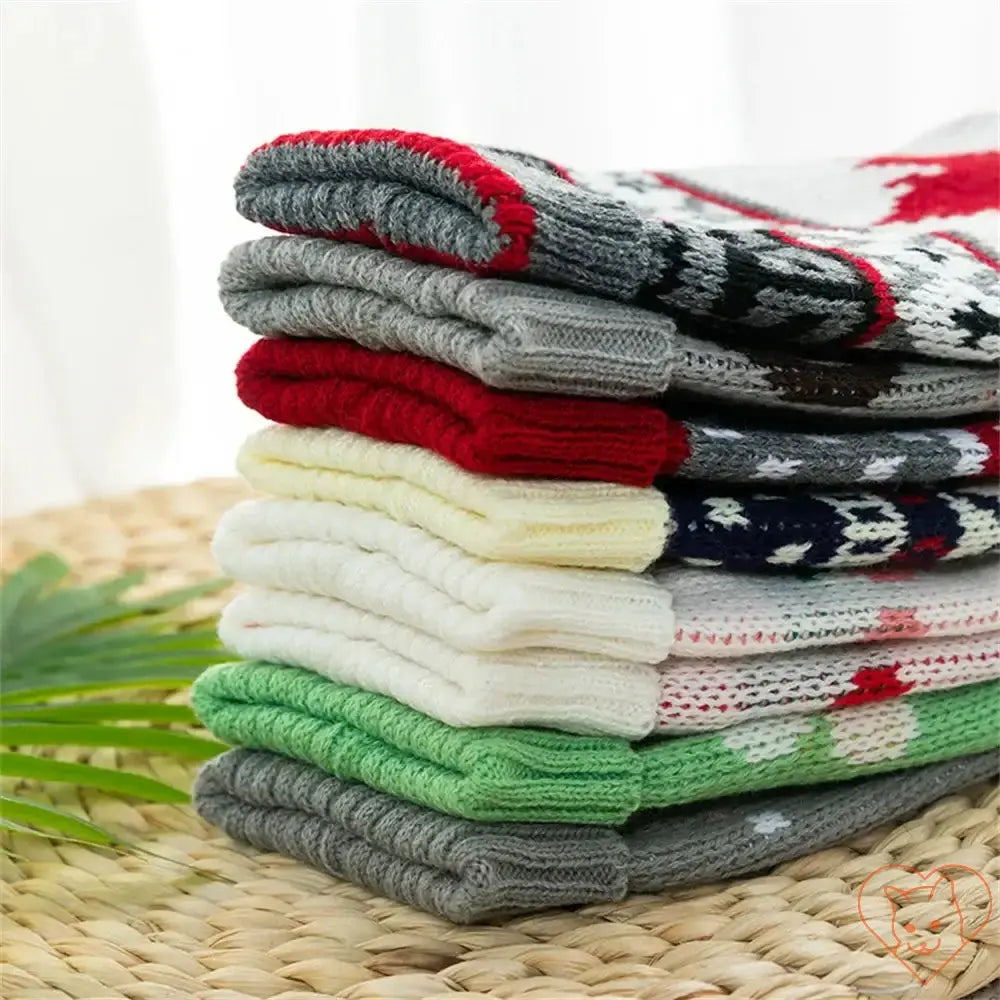 Stack of colorful winter cat sweaters in red, gray, white, and green on a woven surface, perfect for cozy feline fashion.