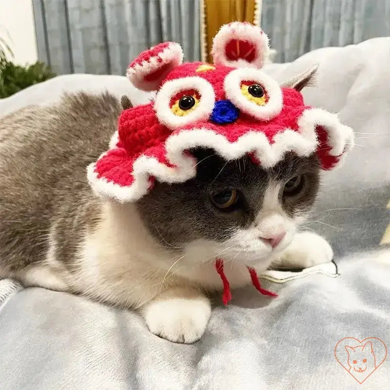 Gray cat wearing a colorful knitted hat with eyes and ears, resting comfortably on a blanket.