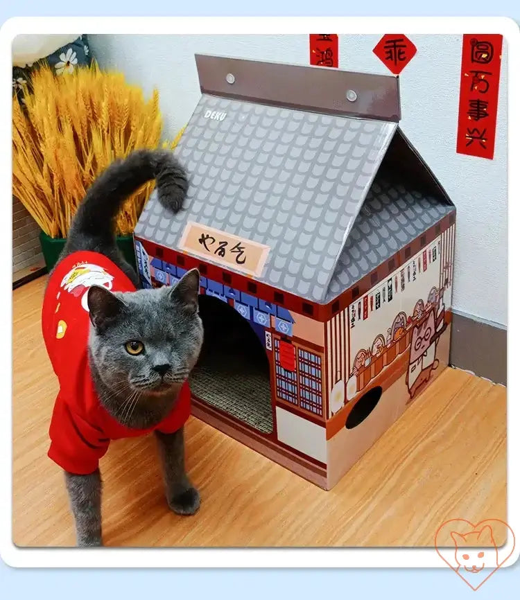 Gray cat in a red shirt beside a colorful cat house designed like a traditional building.