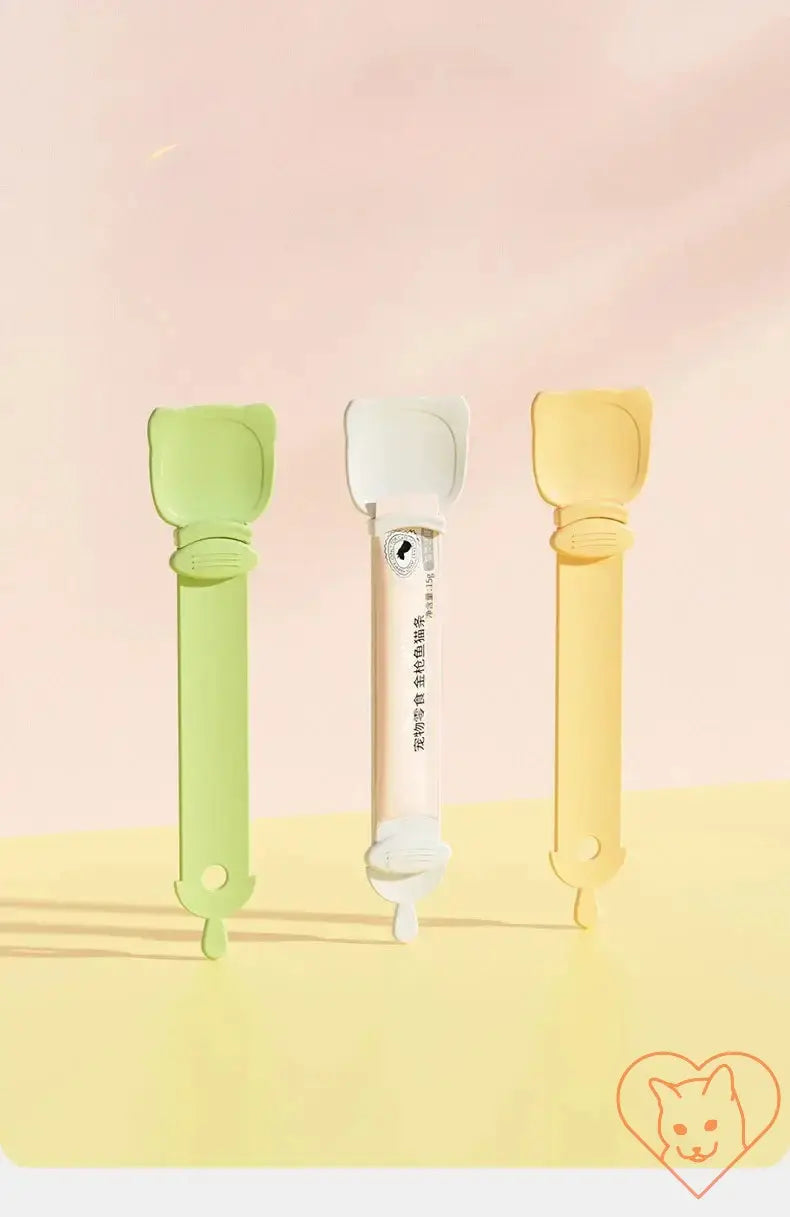 Three multifunctional cat feeding spoons in green, white, and yellow for wet food and treats on a pastel background.